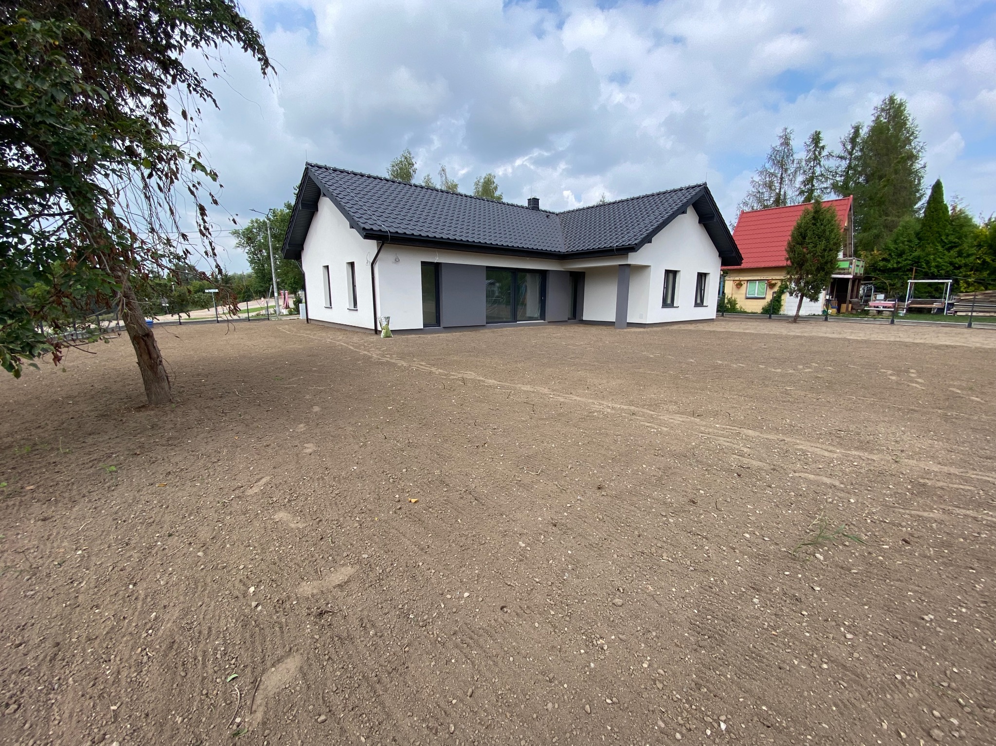 Nowoczesny, parterowy dom z ciemną dachówką i szarymi elementami elewacji, otoczony świeżo wyrównanym terenem pod trawnik.