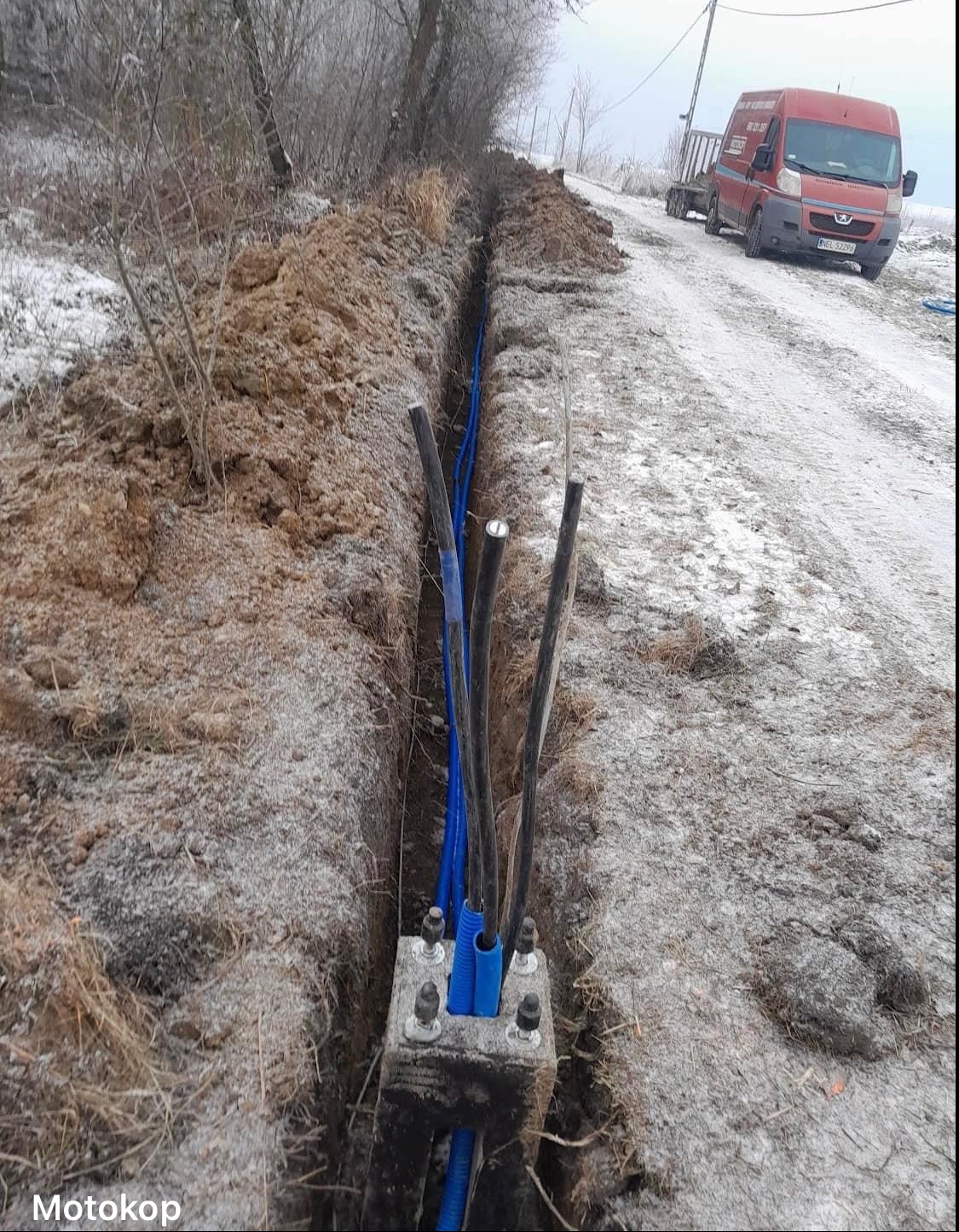 Wykop z ułożonymi kablami elektrycznymi w niebieskich rurach ochronnych, widoczna skrzynka betonowa z mocowaniami, w tle pojazd dostawczy na ośnieżonej drodze.