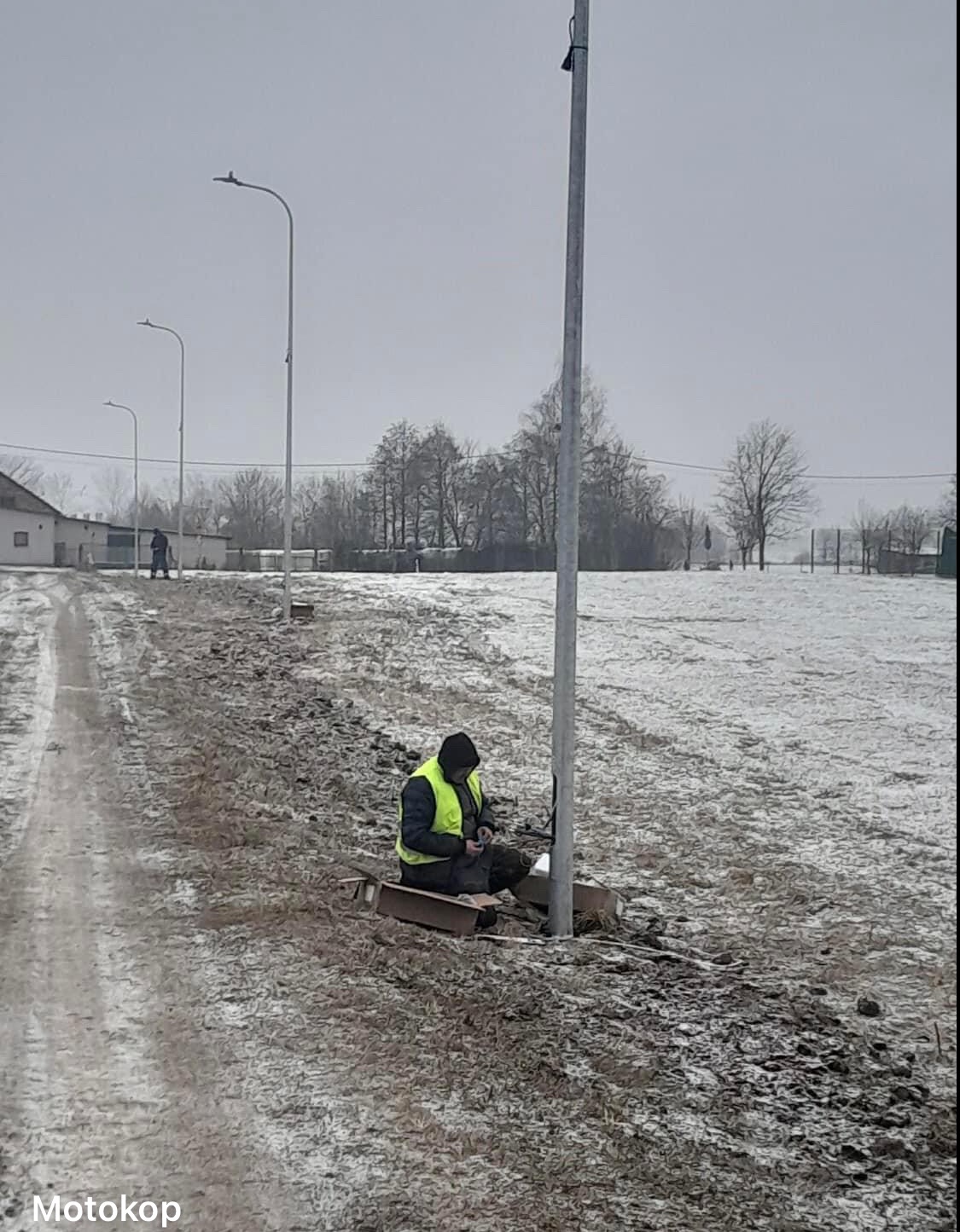 Elektryk w żółtej kamizelce łączy przewody u podstawy słupa oświetleniowego w zimowej scenerii, z widocznymi kolejnymi słupami w oddali i zaśnieżonym polem w tle.