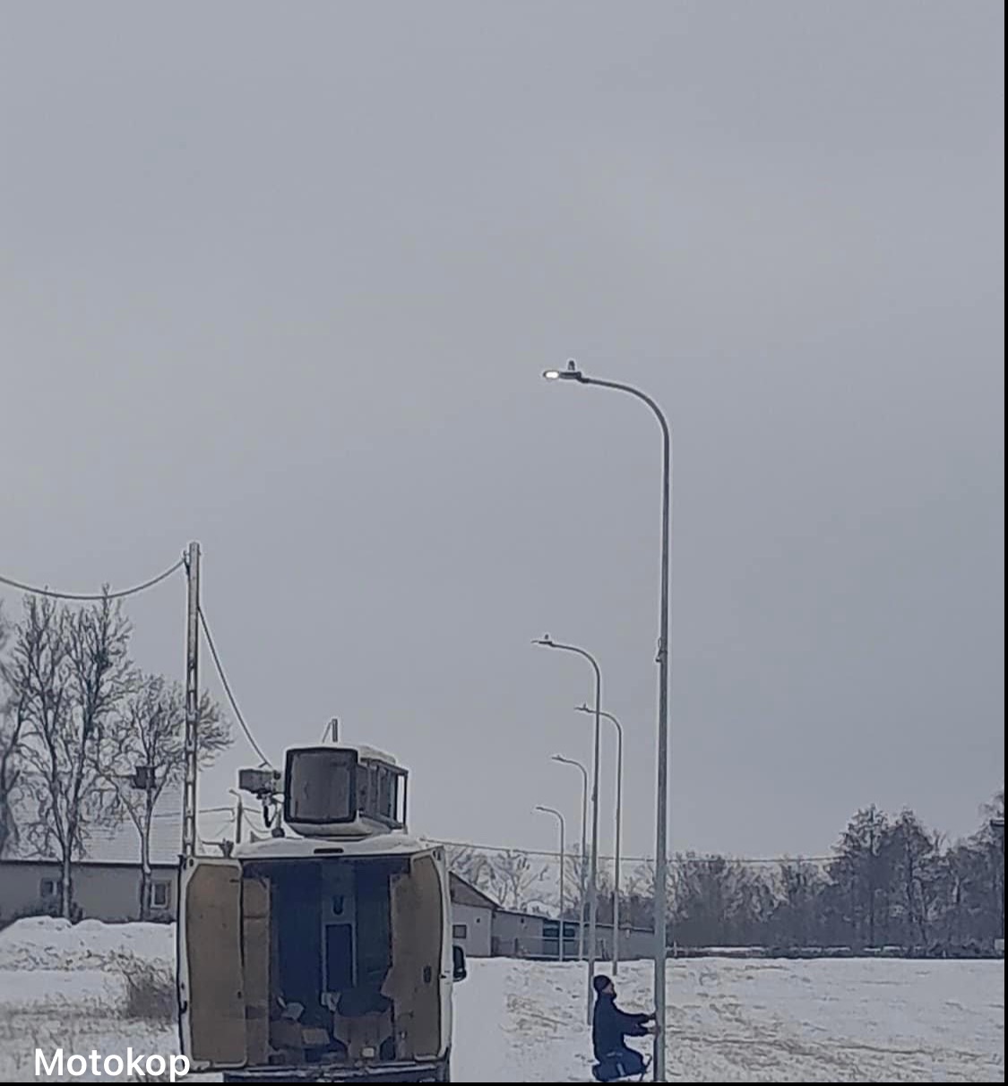 Pracownik w zimowym krajobrazie, montujący oświetlenie uliczne, z otwartym busem serwisowym w tle.