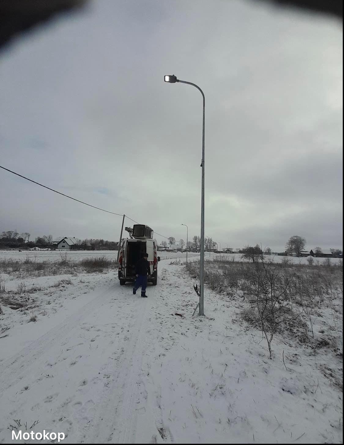 Montaż oświetlenia ulicznego w zimowej scenerii: pracownik w oddali przy furgonetce, świeżo postawiona latarnia z włączonym światłem na zasypanym śniegiem terenie.
