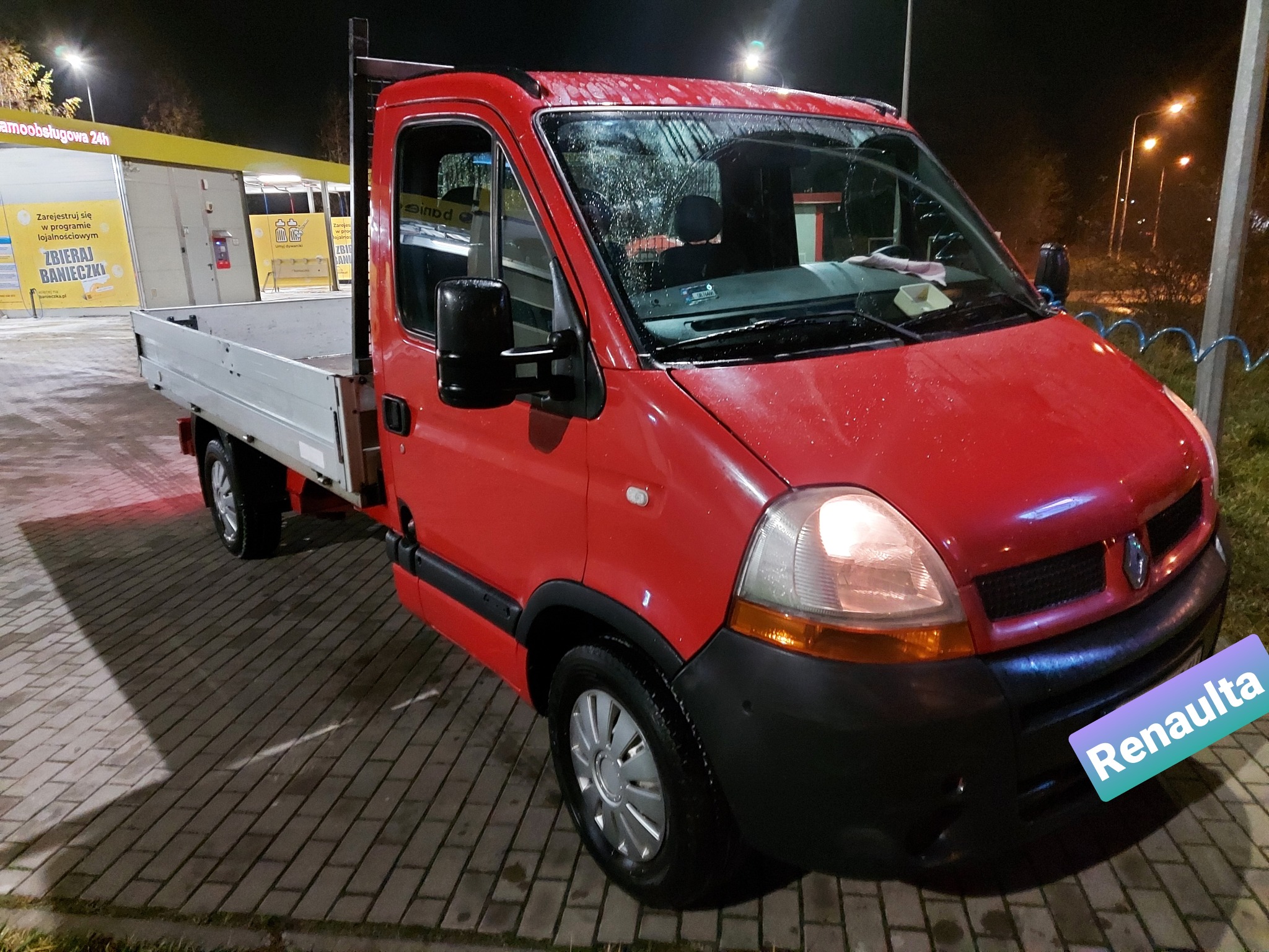 Czerwony samochód dostawczy Renault z otwartą skrzynią ładunkową, zaparkowany nocą przy automatycznej myjni samochodowej.