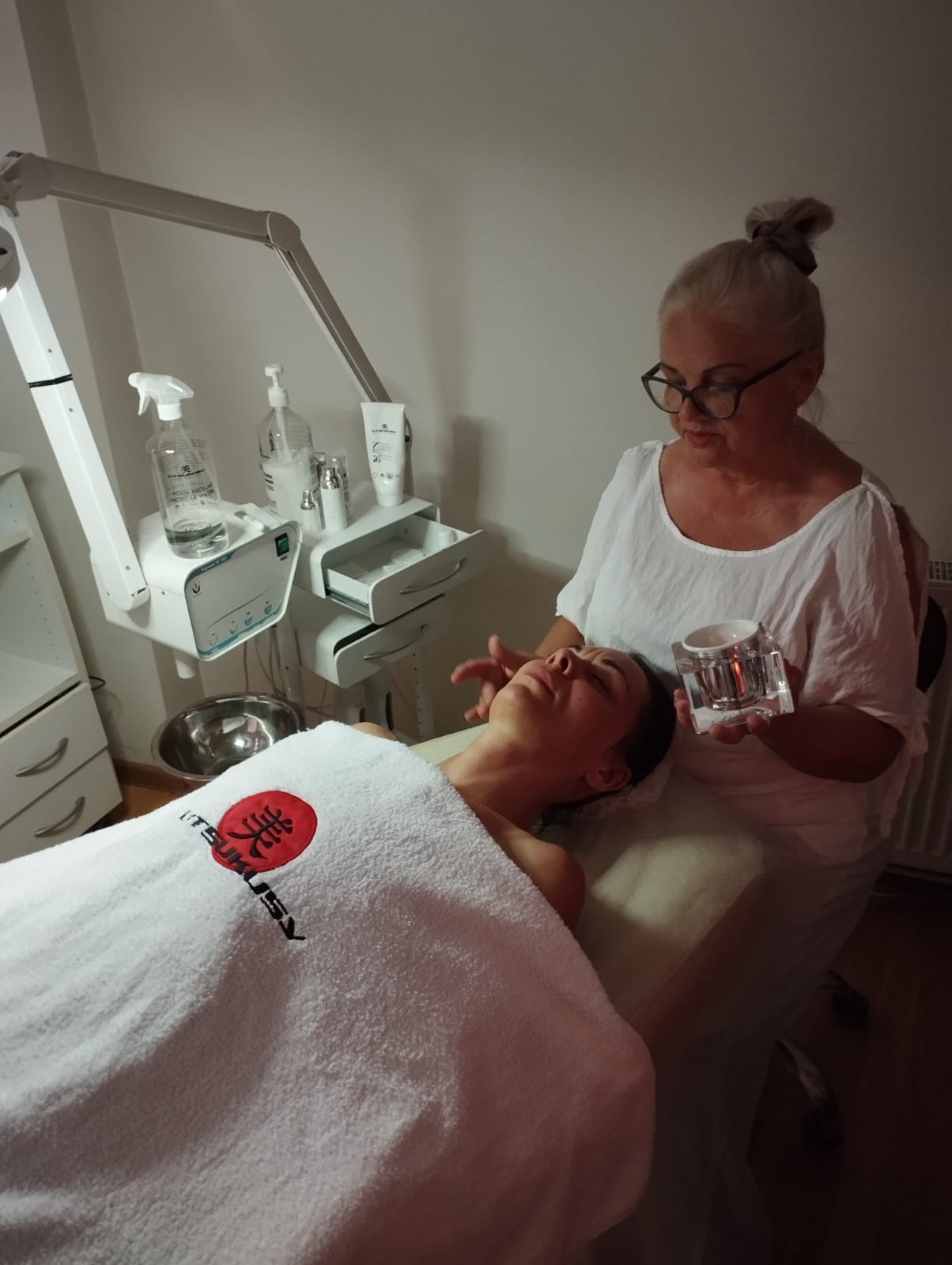 Kosmetyczka wykonuje masaż twarzy klientce leżącej na leżance, na której leży biały ręcznik z logo firmy kosmetycznej, a w tle widać sprzęt kosmetyczny i kosmetyki.