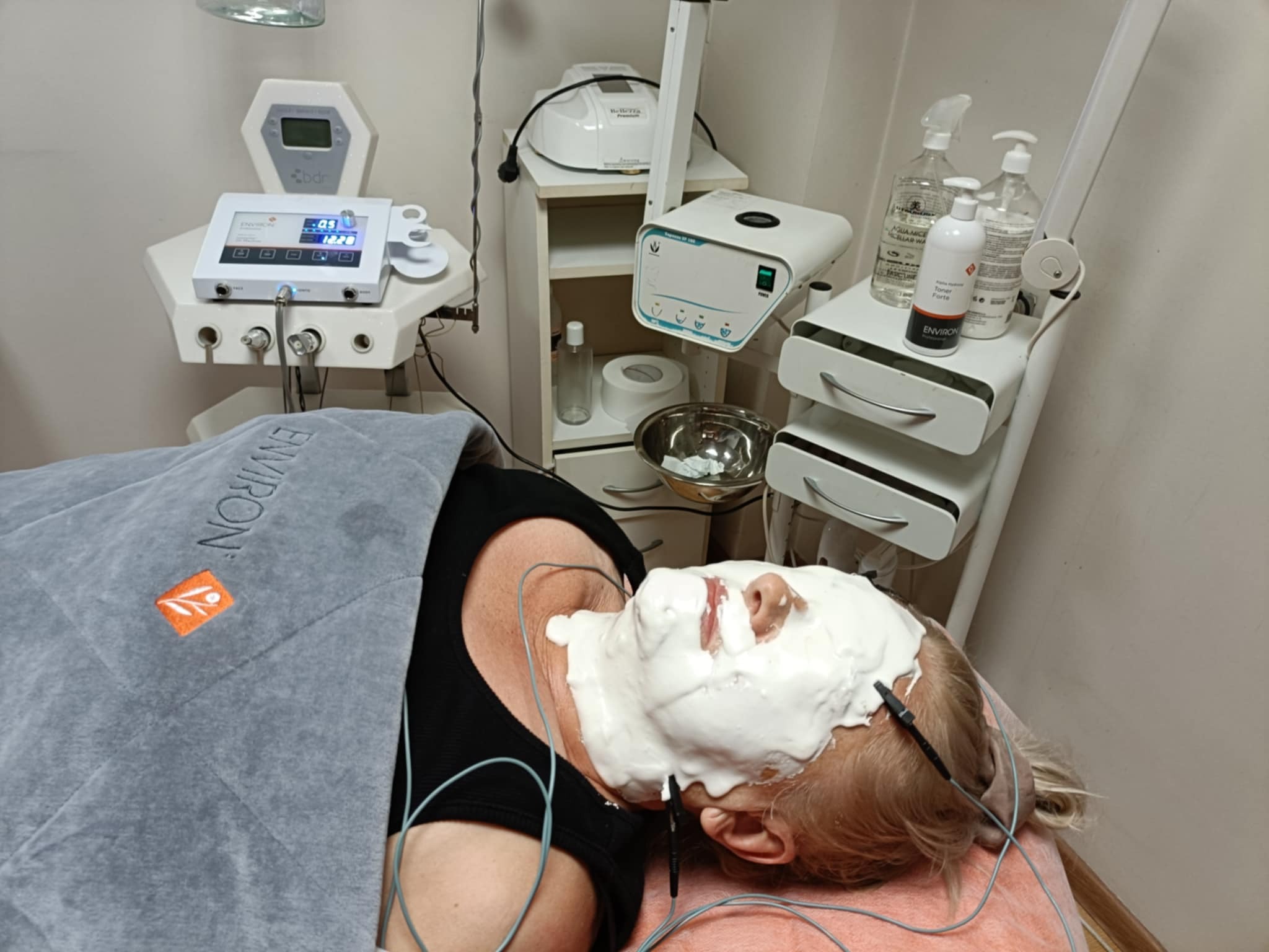 Klientka leżąca na leżance z maską na twarzy i podłączonymi elektrodami, obok widoczne urządzenia kosmetyczne i preparaty w salonie.