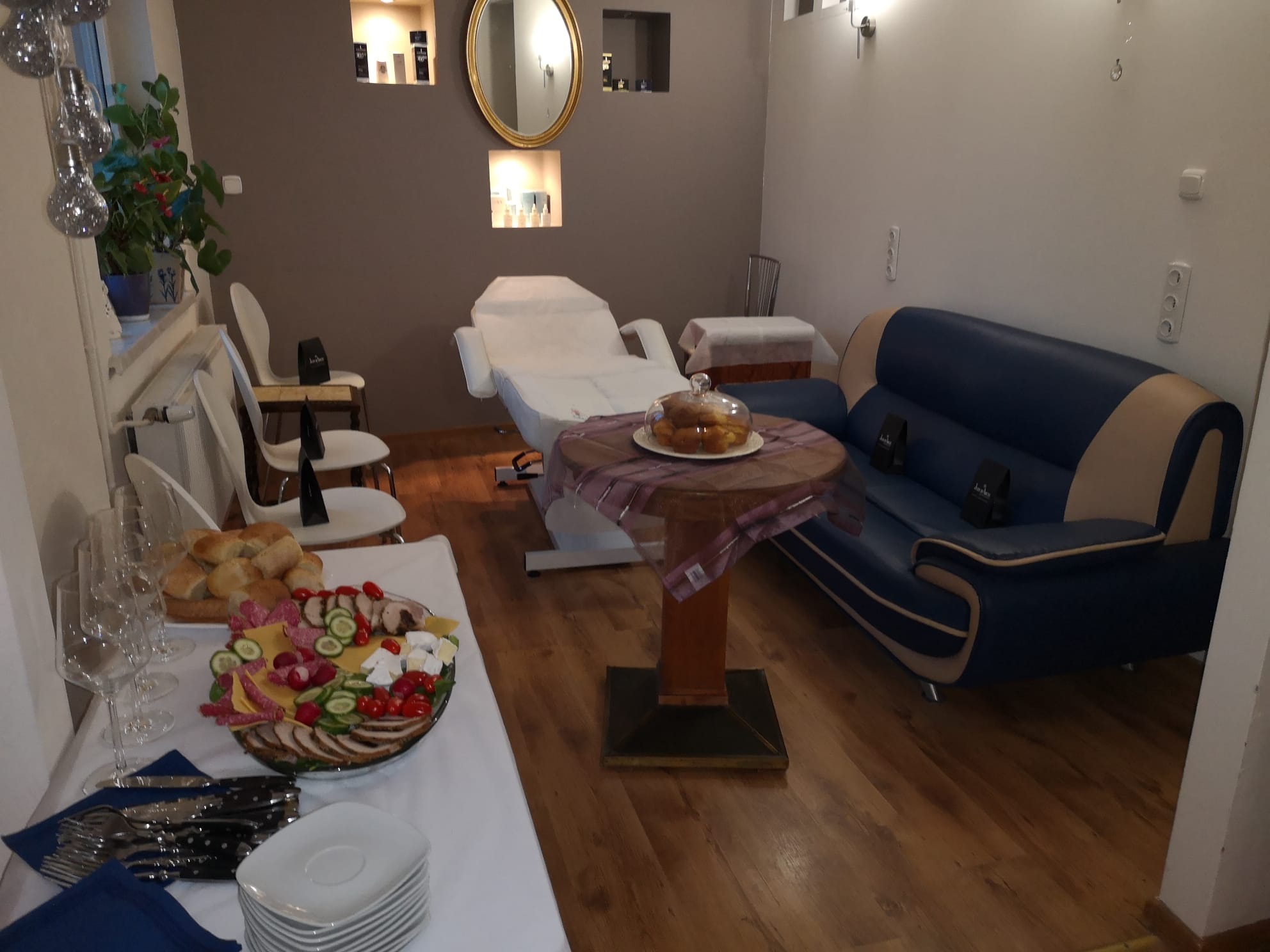 Wnętrze gabinetu kosmetycznego z przygotowanym poczęstunkiem: stół z przekąskami, ciasto pod kloszem, niebieska sofa i białe fotele, w tle łóżko kosmetyczne.