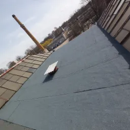 Widok z góry na płaski dach pokryty papą, z kominkiem wentylacyjnym z daszkiem, w tle komin fabryczny i budynki.