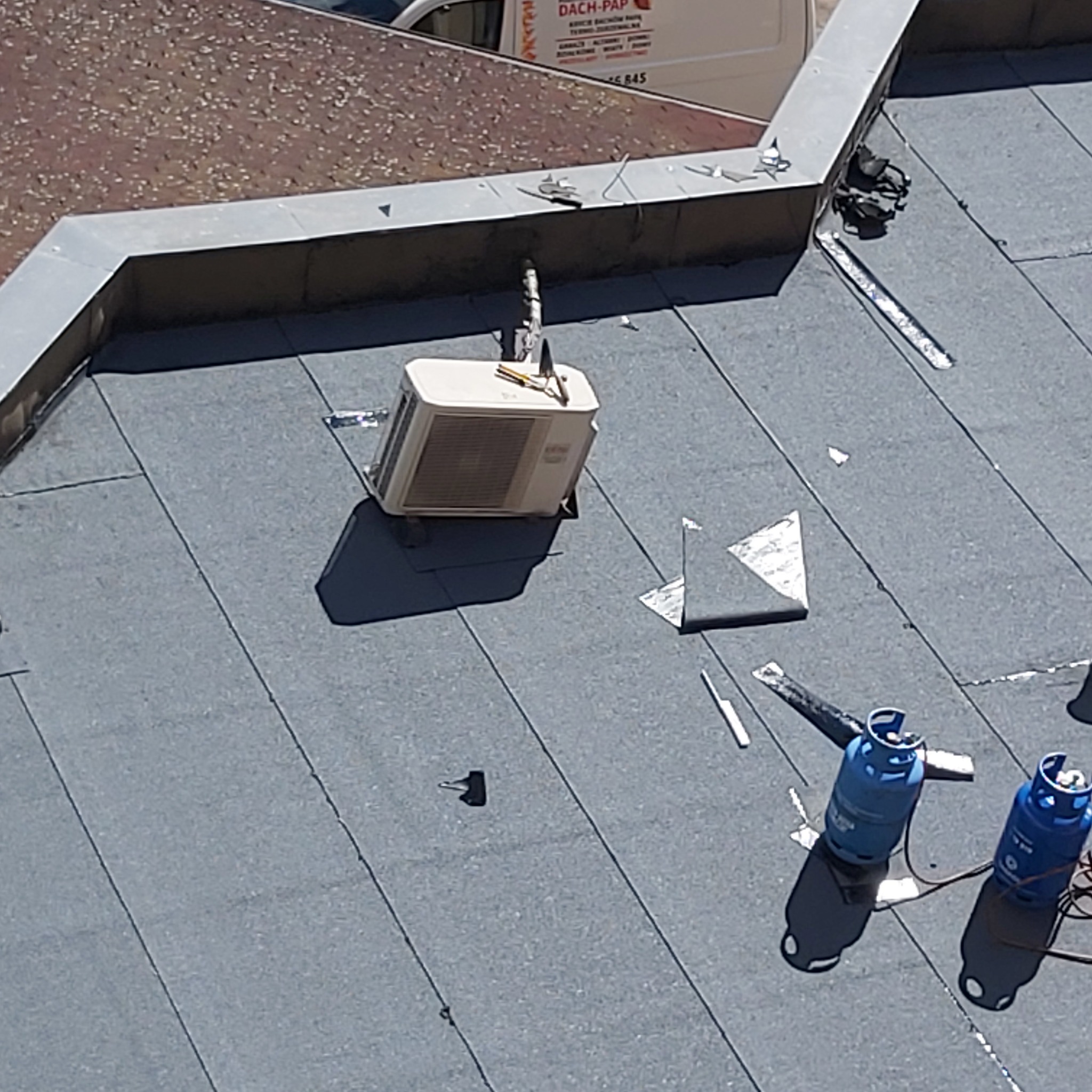 Widok z góry na płaski dach pokryty papą, z widocznymi butlami gazowymi, klimatyzatorem i narzędziami dekarskimi, sugerujący prace związane z uszczelnianiem lub naprawą.