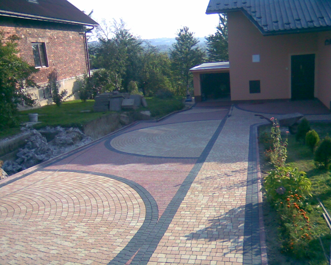 Ułożona kostka brukowa w odcieniach beżu i czerwieni tworząca wzór wachlarza na podjeździe, oddzielona ciemniejszym pasem kostki, widok na domy i zieleń w tle.