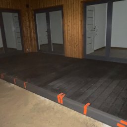 Green Building - Ciemny taras z desek, częściowo oświetlony, z pomarańczowymi oznaczeniami na krawędziach, przylegający do budynku z drewnianą elewacją i trzema parami drzwi balkonowych.