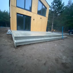 Green Building - Nowoczesny dom modułowy z jasną drewnianą elewacją i szarym tarasem z dwoma stopniami, widok z poziomu gruntu na tle zalesionego terenu.