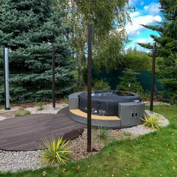 Green Building - Nowoczesna, okrągła wanna spa otoczona drewnianym tarasem i ozdobnym żwirem w ogrodzie z iglakami i prysznicem na zewnątrz.