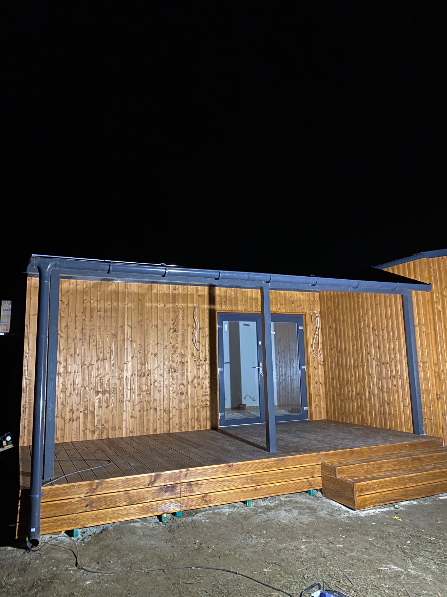 Nowoczesny dom modułowy z drewnianą elewacją i tarasem, widoczne szare ramy okienne i drzwi, oświetlenie nocne.