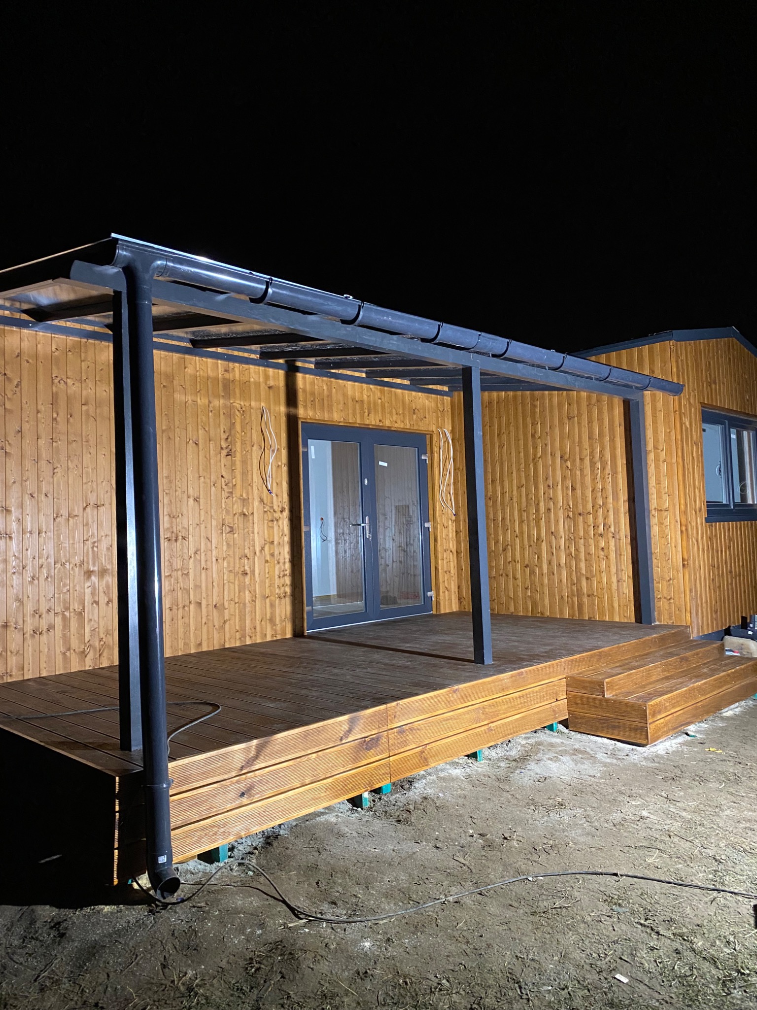 Nowoczesny dom modułowy z drewnianą elewacją, tarasem z desek i metalową pergolą w nocnym oświetleniu.