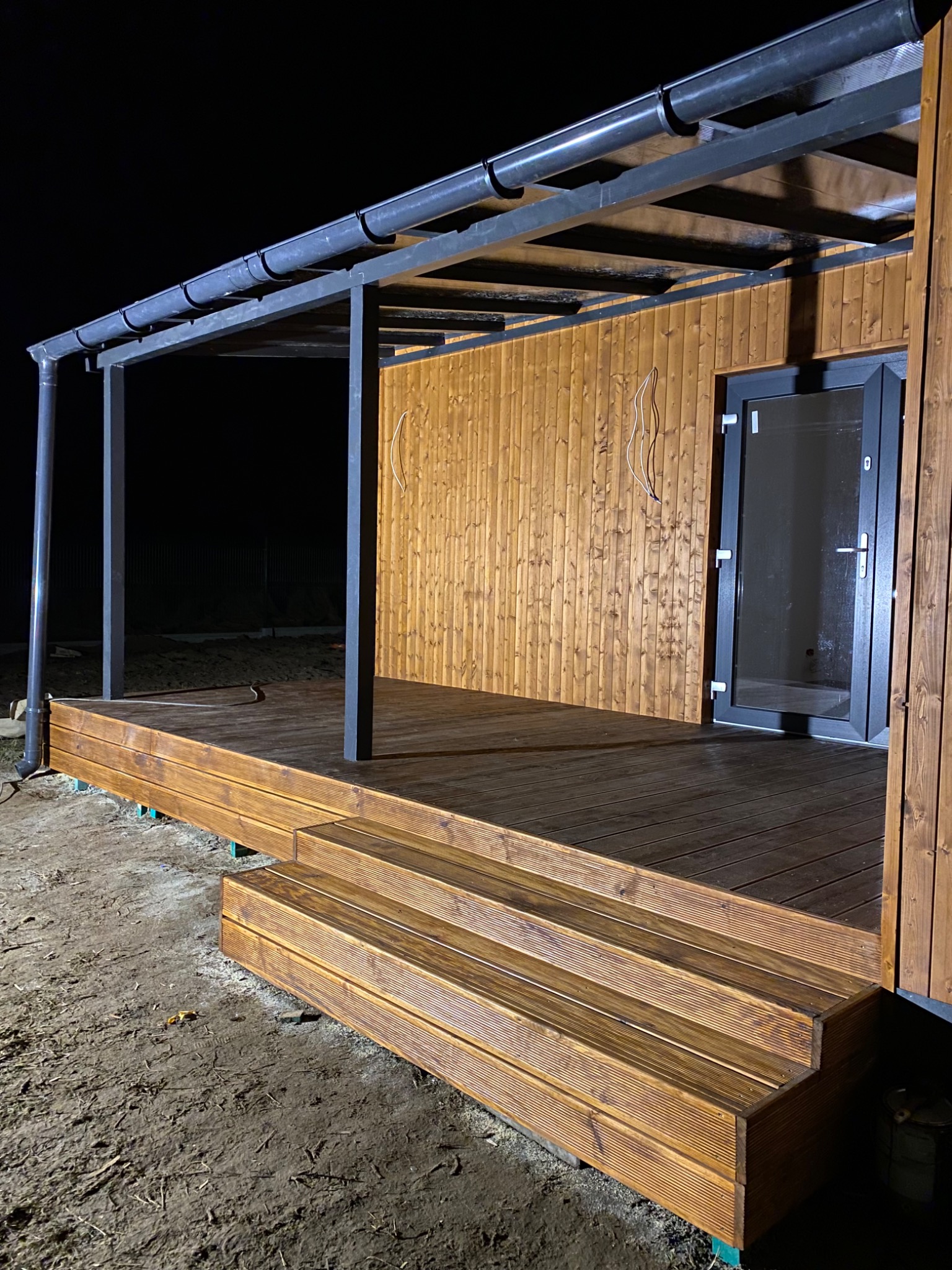 Zadaszony taras z drewnianą podłogą i schodami w Warszawie, oświetlony nocą, widoczny fragment elewacji domu z drewnianą boazerią i szarą stolarką okienną.