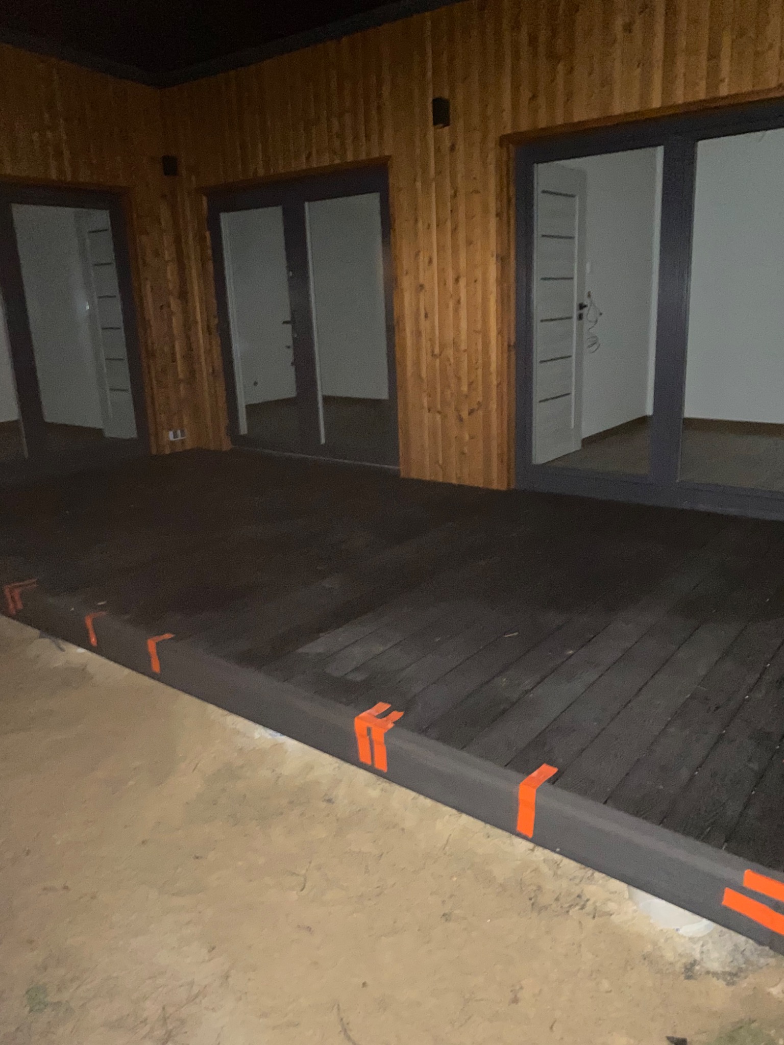 Ciemny taras z desek, częściowo oświetlony, z pomarańczowymi oznaczeniami na krawędziach, przylegający do budynku z drewnianą elewacją i trzema parami drzwi balkonowych.