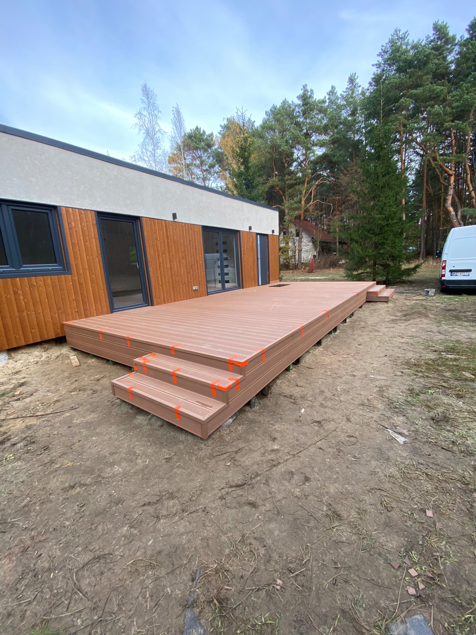 Nowoczesny dom modułowy z brązową elewacją i tarasem kompozytowym z trzema schodkami, zabezpieczony pomarańczową taśmą, na tle lasu i pochmurnego nieba.