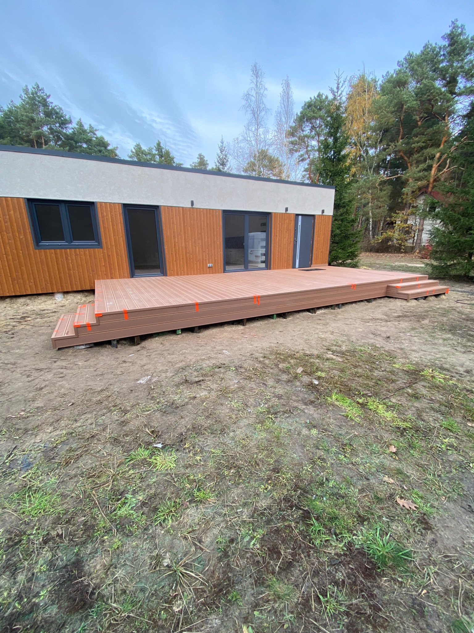 Nowoczesny dom modułowy z tarasem z desek kompozytowych w kolorze brązowym, z widocznymi schodkami i pomarańczowymi taśmami zabezpieczającymi, otoczony trawnikiem i drzewami w pochmurny dzień.