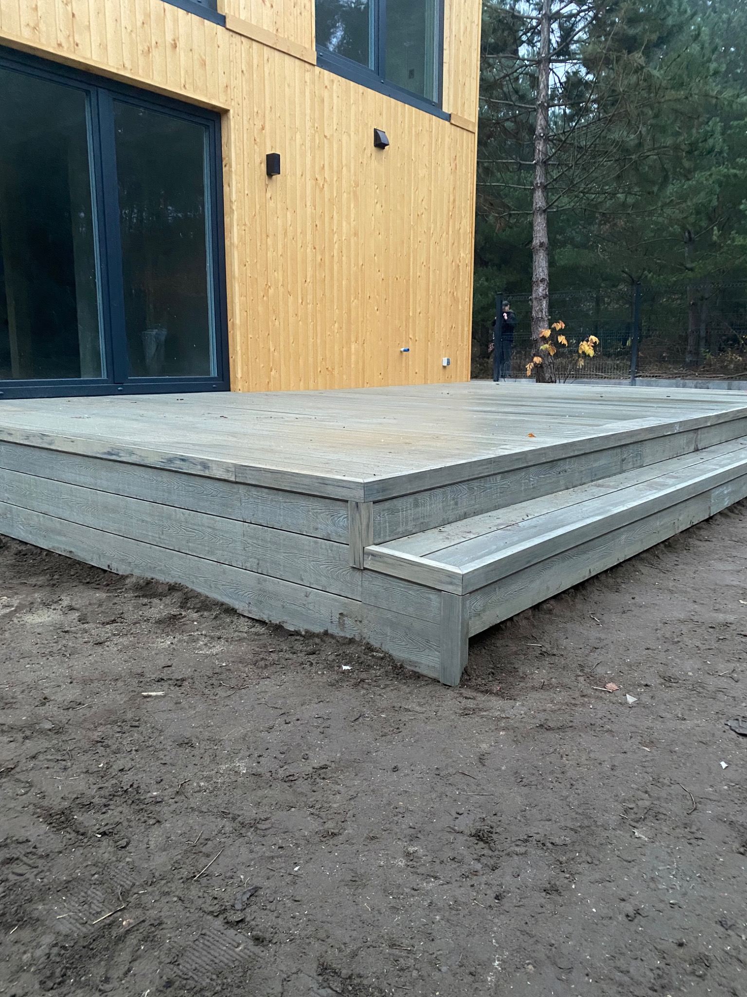 Nowoczesny taras z kompozytu w odcieniach szarości, z niskimi schodami, przylegający do budynku o jasnej, drewnianej elewacji. W tle widoczne drzewa iglaste i osoba stojąca przy ogrodzeniu.