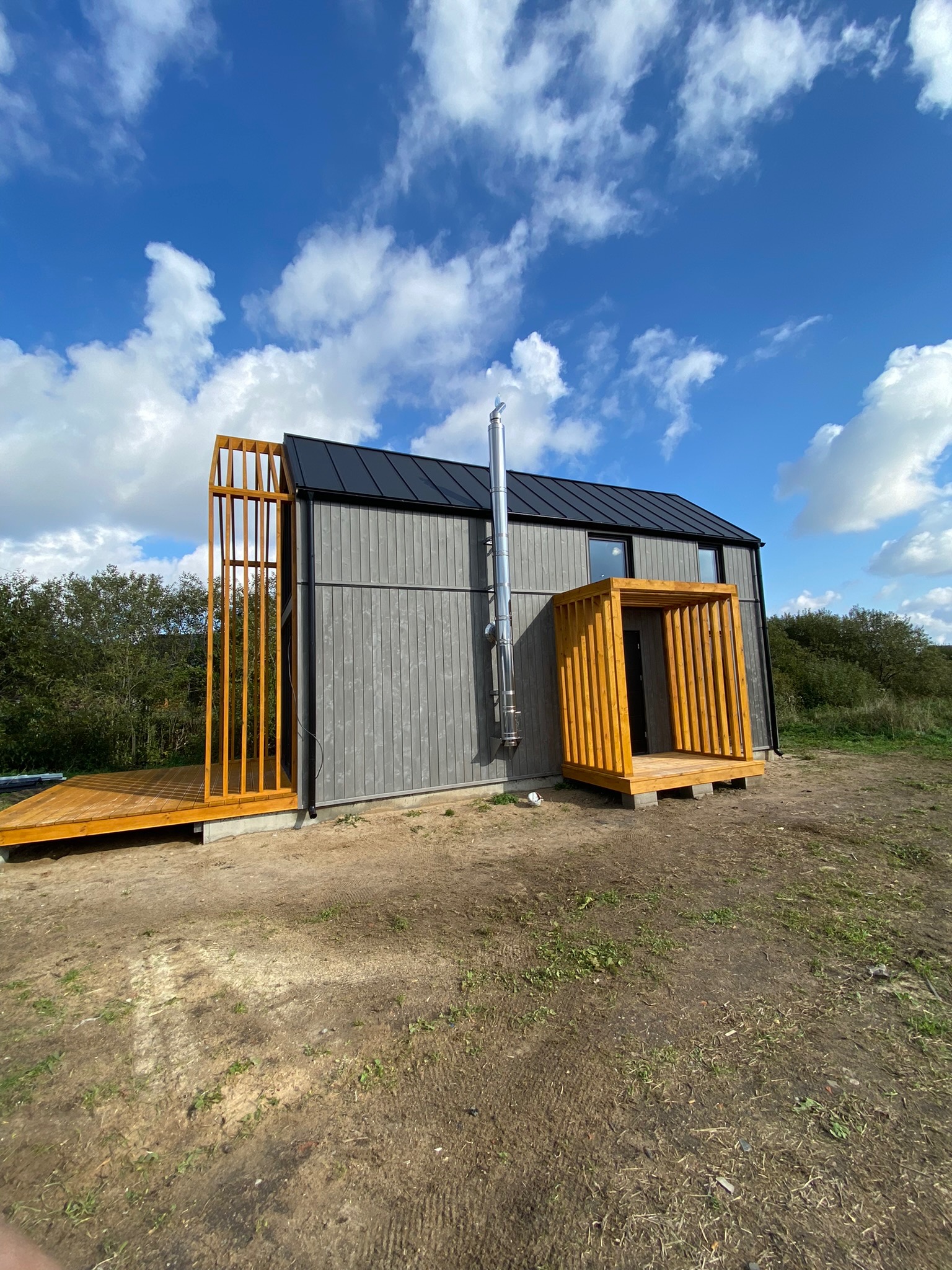 Nowoczesny dom modułowy z szarej elewacji z drewna, z drewnianym tarasem i wejściem, komin ze stali nierdzewnej, na tle błękitnego nieba z chmurami.