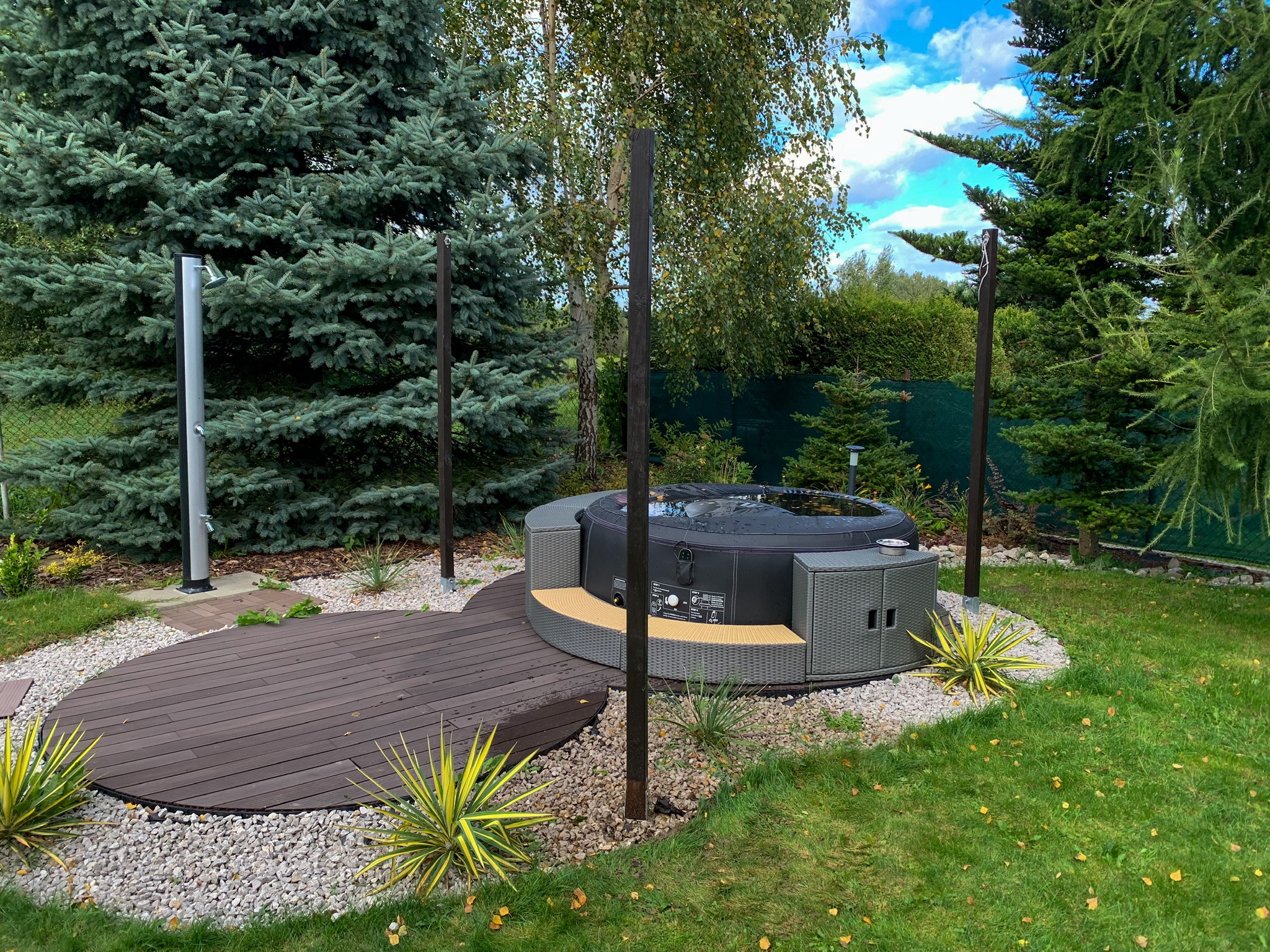 Nowoczesna, okrągła wanna spa otoczona drewnianym tarasem i ozdobnym żwirem w ogrodzie z iglakami i prysznicem na zewnątrz.