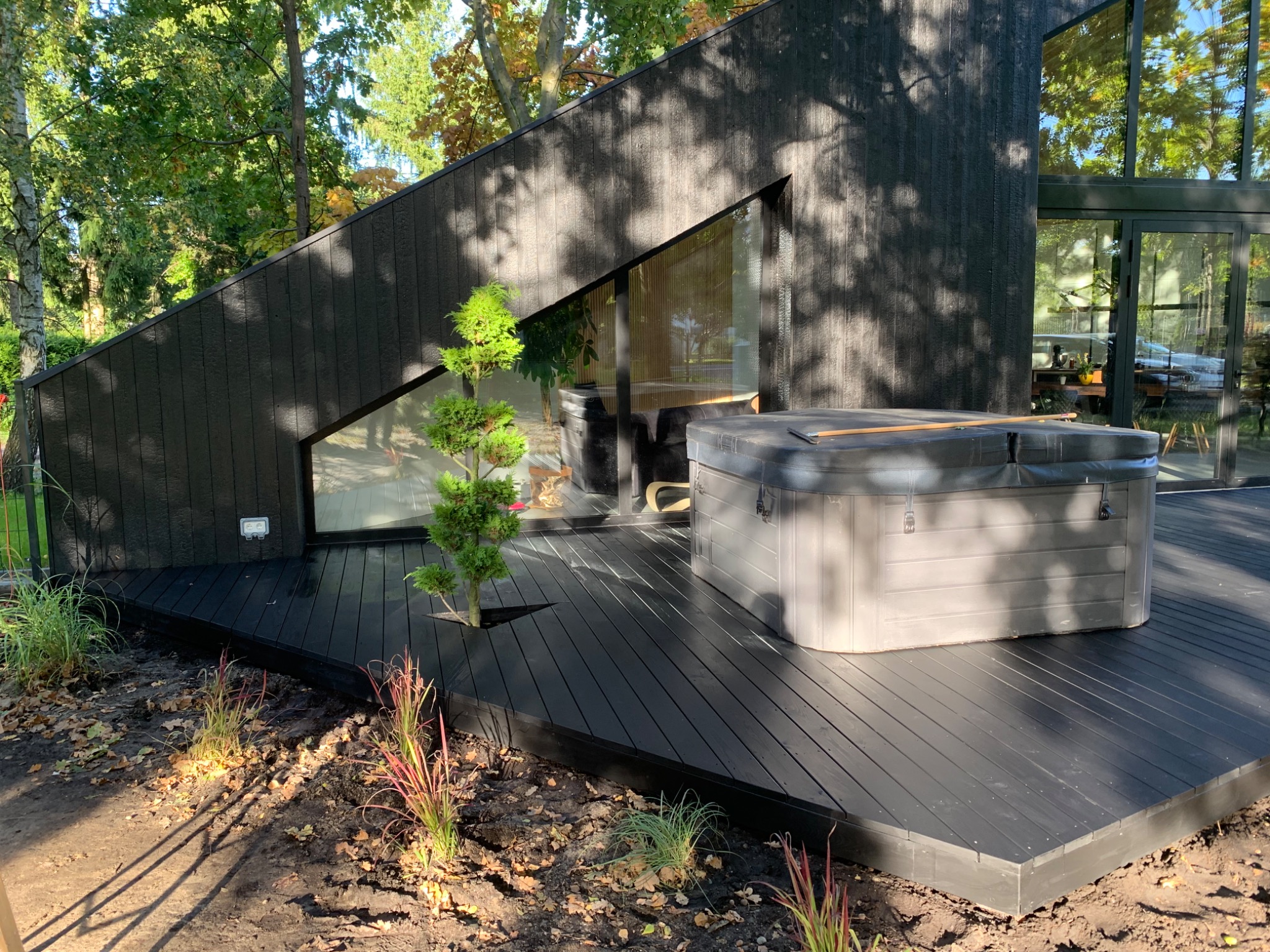 Nowoczesny taras z ciemnego drewna przylegający do budynku o nietypowej architekturze, z jacuzzi i małym drzewkiem zasadzonym bezpośrednio w deski tarasu.