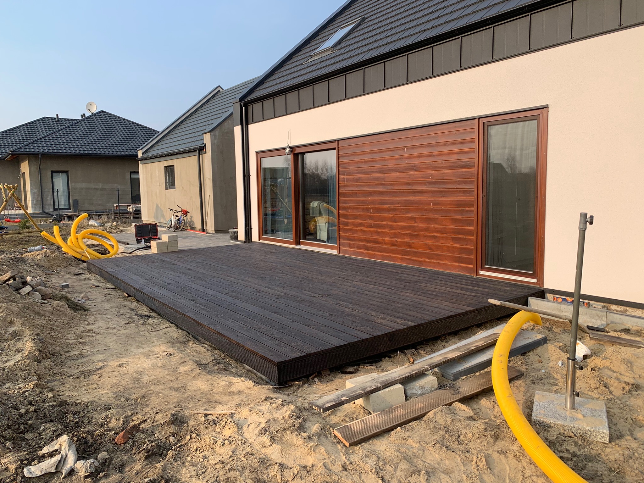 Ciemnobrązowy taras drewniany w trakcie budowy przy nowoczesnym domu z drewnianą elewacją i żółtym wężem wentylacyjnym na piaszczystym podłożu.