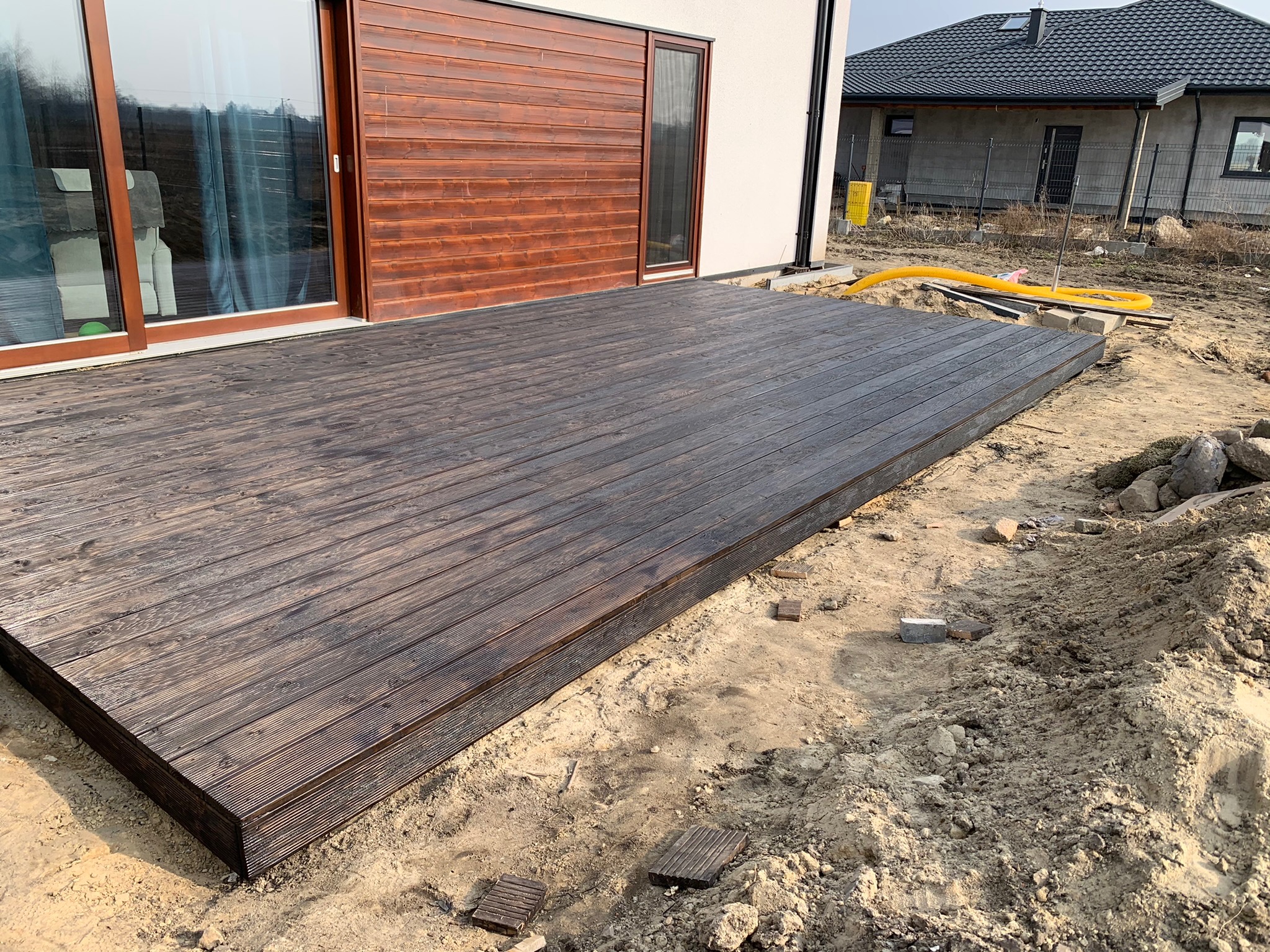 Nowoczesny taras z ciemnego drewna przylegający do budynku z drewnianą elewacją, widoczne przygotowane podłoże z piasku i kamieni, w tle dom w stanie surowym.