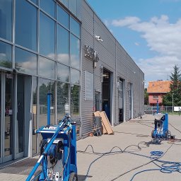 MaR-GaRet firma sprzątająco porządkowa Małgorzata Kasprzykowska - Profesjonalne mycie elewacji budynku serwisowego z użyciem specjalistycznego sprzętu ciśnieniowego. Widoczne niebieskie maszyny i węże na tle szarej elewacji.