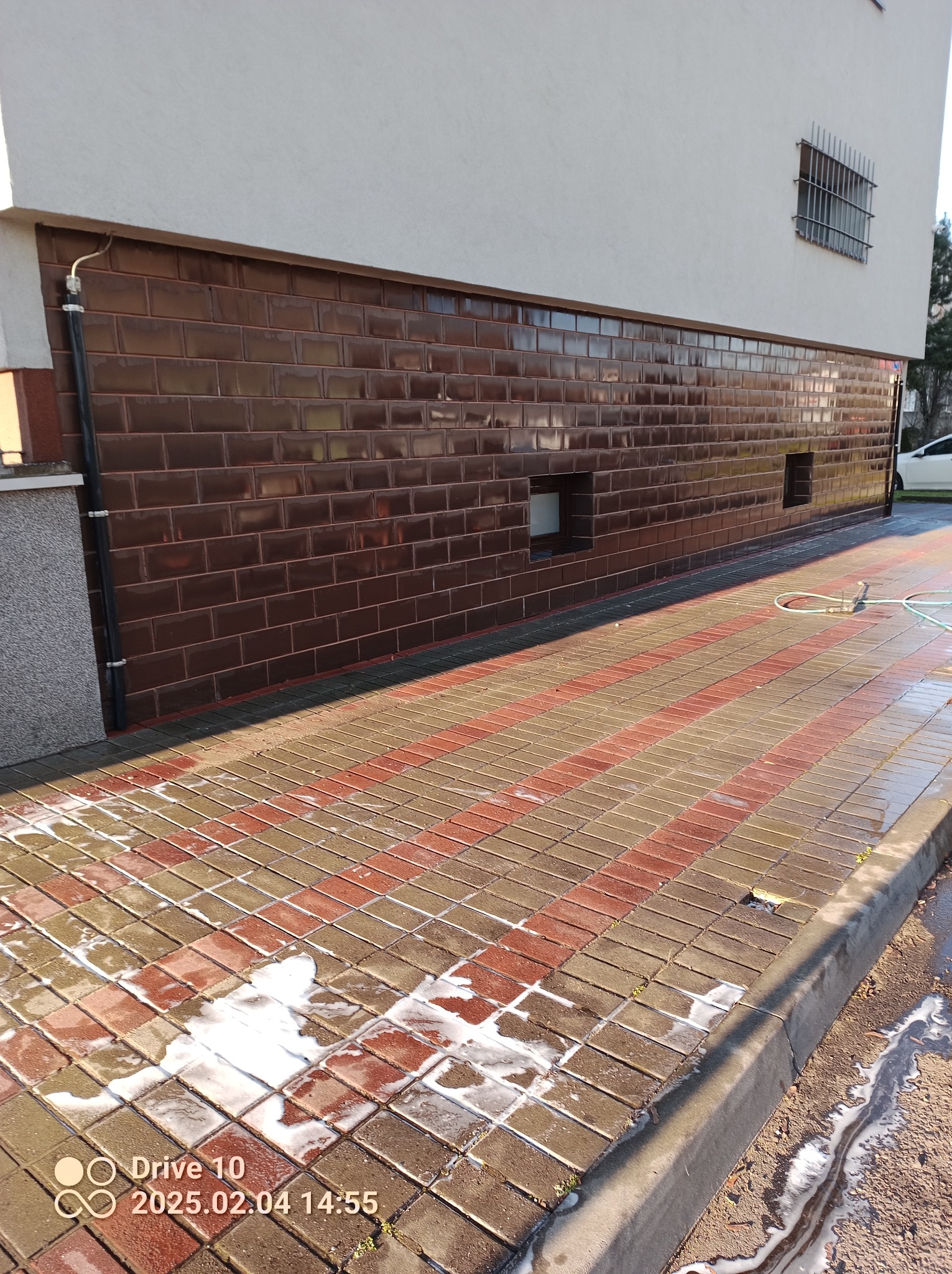 Częściowo umyta kostka brukowa przed budynkiem z brązową, błyszczącą elewacją z płytek, widoczny wąż ogrodowy i resztki piany czyszczącej.