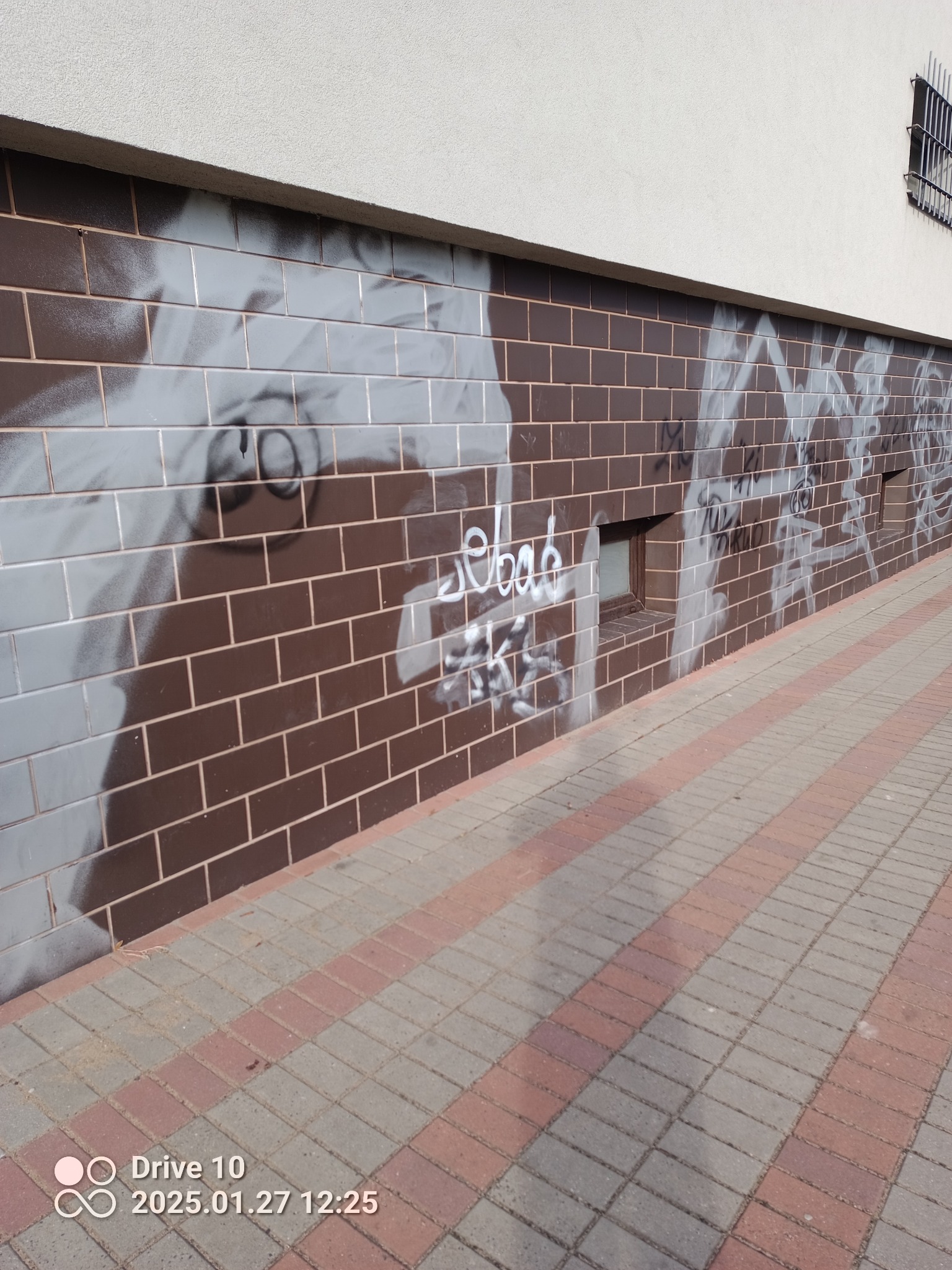 Elewacja budynku z brązowych płytek częściowo pokryta graffiti, widoczne małe okienka wentylacyjne i chodnik z kostki brukowej.