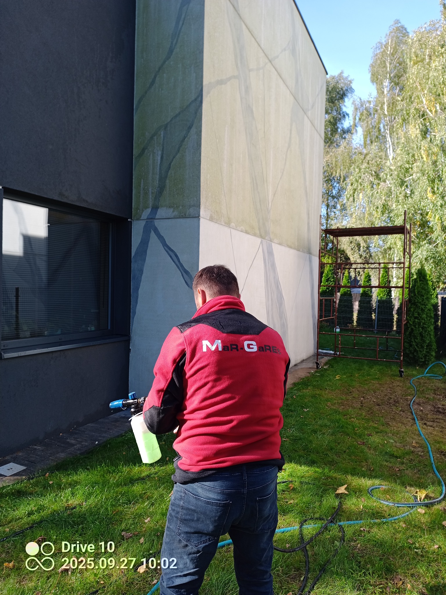 Pracownik w czerwonej bluzie czyści elewację budynku z zanieczyszczeń za pomocą myjki ciśnieniowej. Widoczne rusztowanie i trawnik.