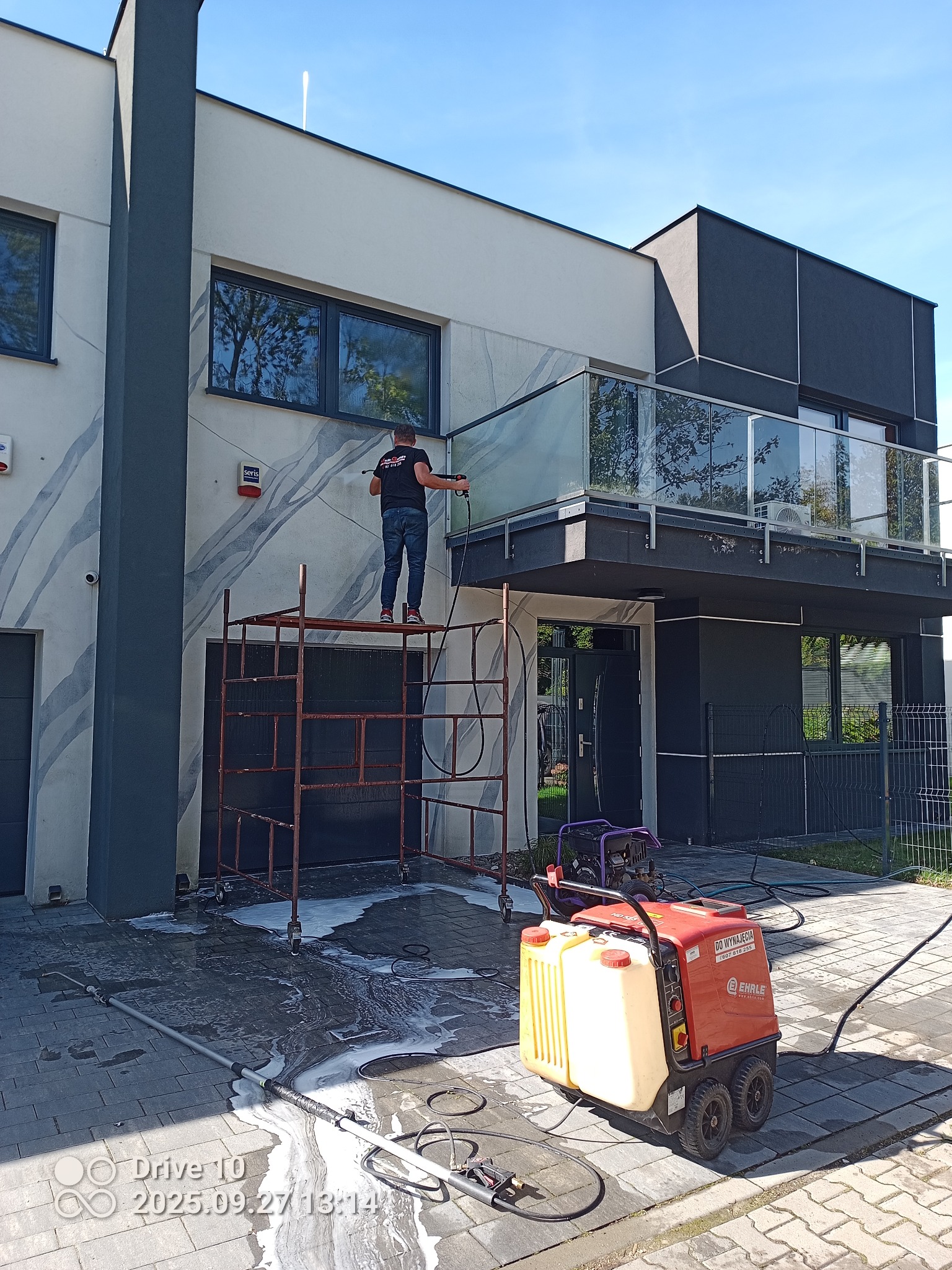 Pracownik na rusztowaniu czyści elewację nowoczesnego domu myjką ciśnieniową. Widoczny sprzęt do mycia i ślady wody z pianą na kostce brukowej. Jasne, słoneczne oświetlenie.
