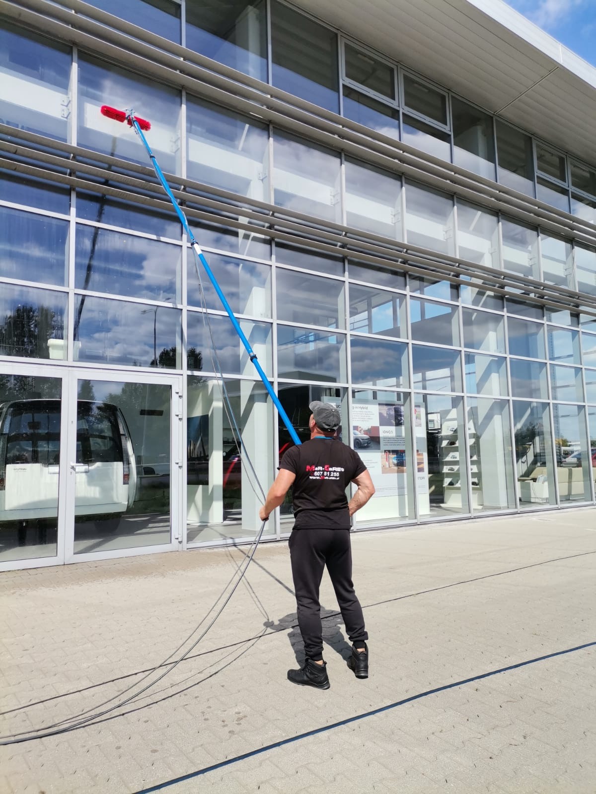 Pracownik w czarnym stroju czyści szklaną fasadę budynku za pomocą długiej, niebieskiej lancy z czerwoną szczotką. Widok z dołu, budynek odbija niebo.