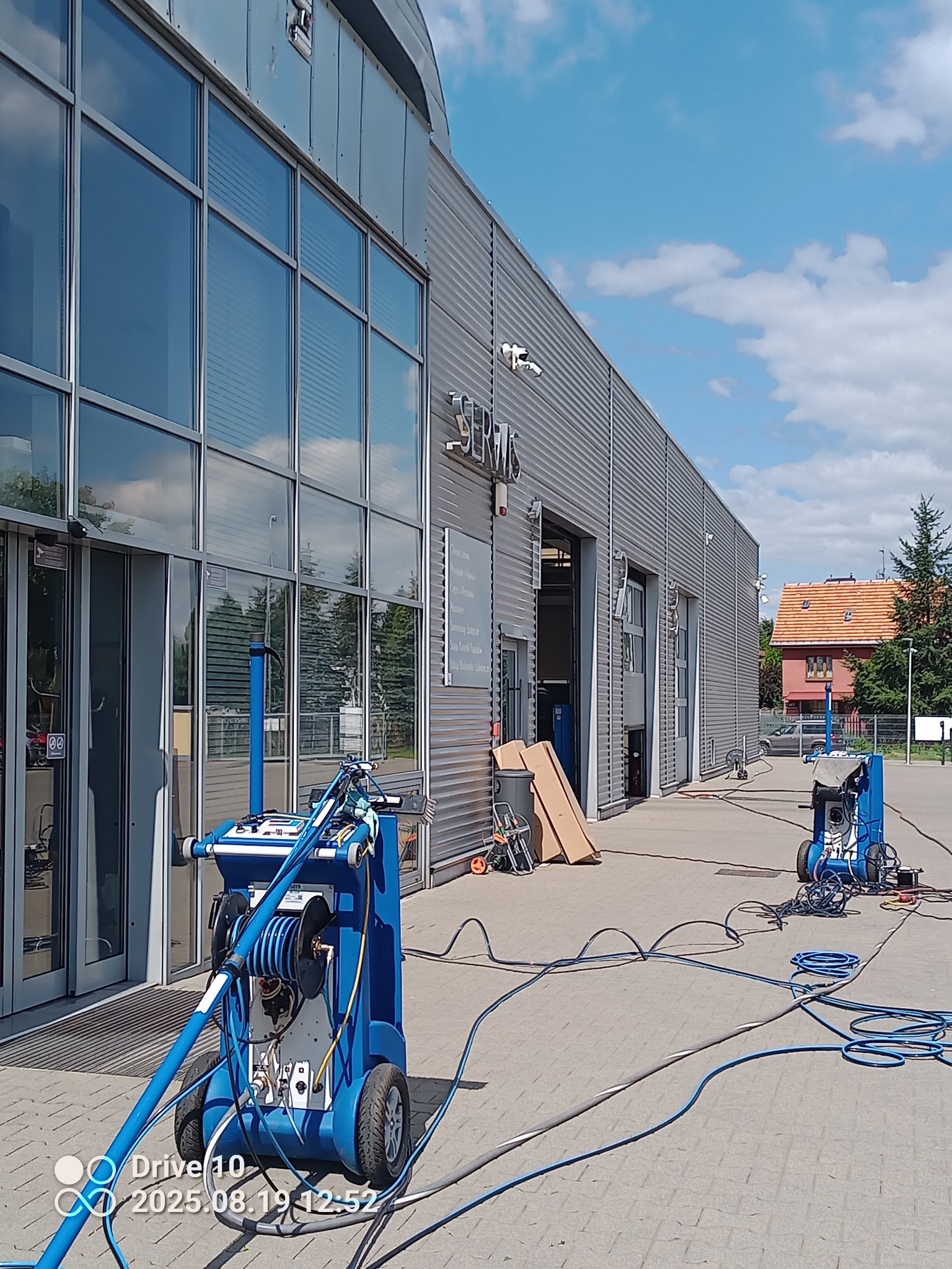 Profesjonalne mycie elewacji budynku serwisowego z użyciem specjalistycznego sprzętu ciśnieniowego. Widoczne niebieskie maszyny i węże na tle szarej elewacji.