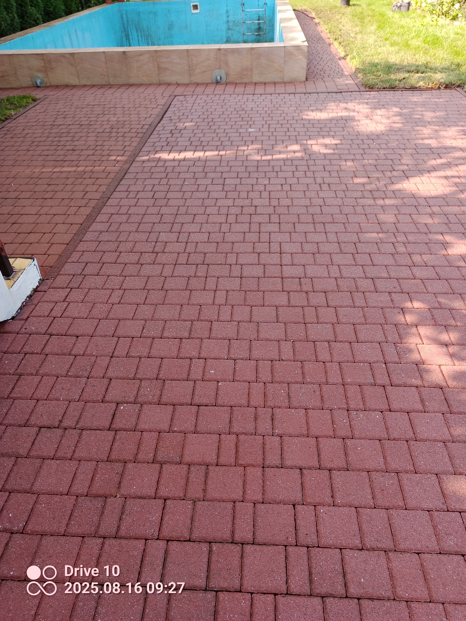 Czerwona kostka brukowa ułożona wokół basenu, widoczne cienie drzew. Regularny wzór kostki, jasne fugi. Ujęcie z góry, podkreślające geometrię i porządek.