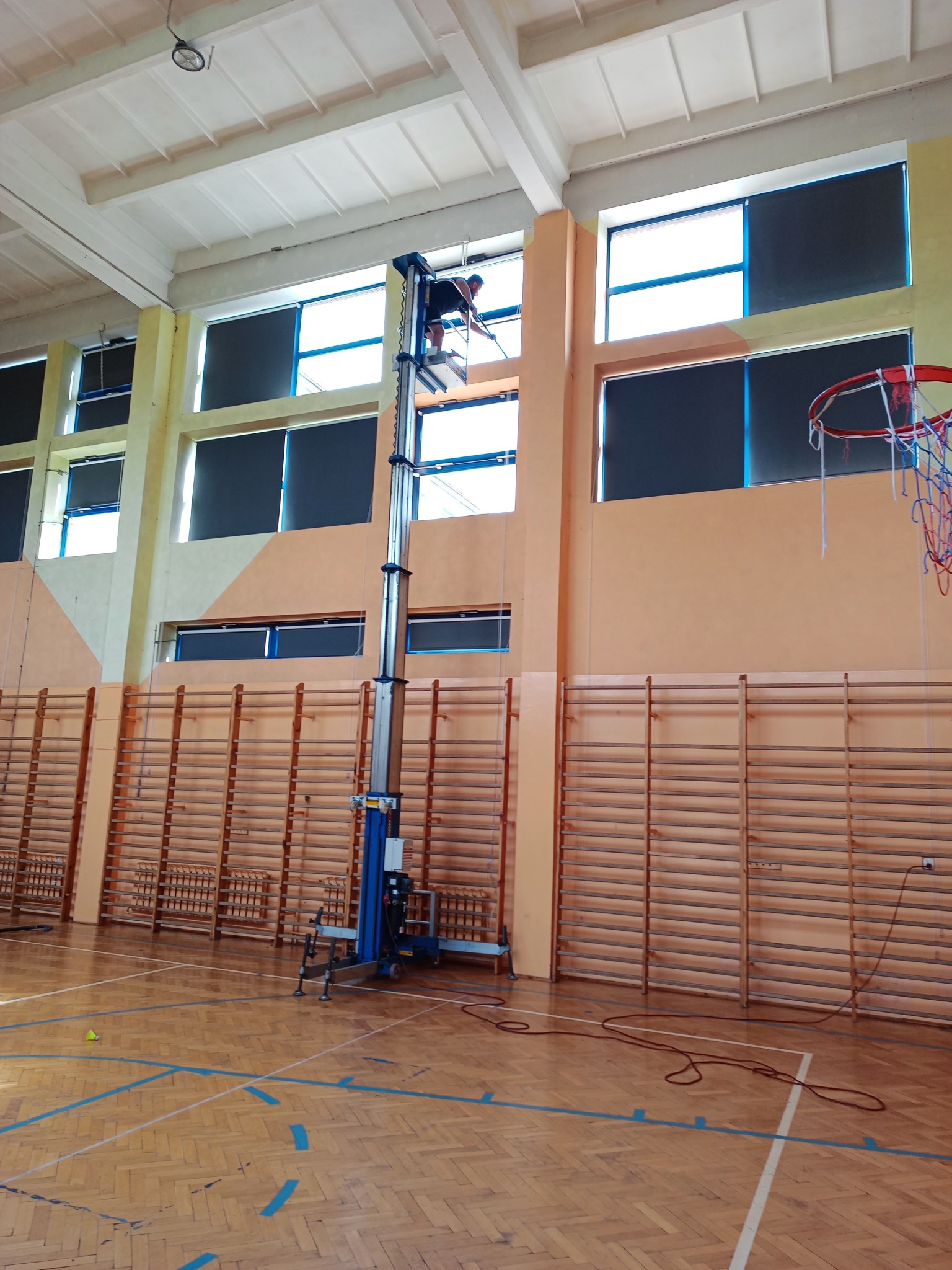 Pracownik na podnośniku czyści okna w hali sportowej. Widoczne drabinki gimnastyczne i kosz do koszykówki. Hala oświetlona naturalnym światłem.