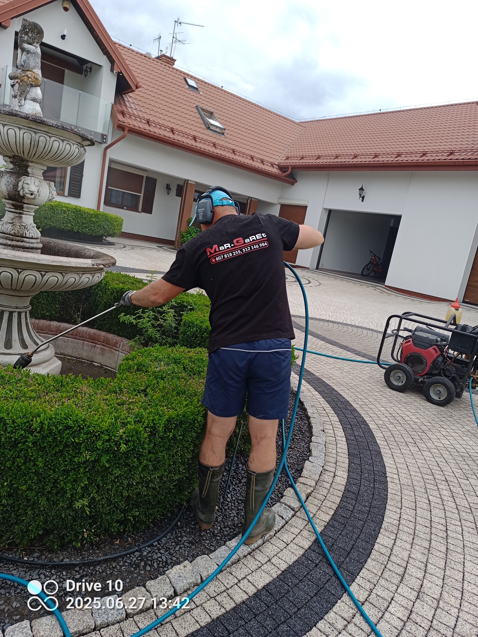 Pracownik w trakcie czyszczenia fontanny myjką ciśnieniową na tle domu z czerwoną dachówką i kostki brukowej. Widoczny sprzęt do mycia ciśnieniowego.