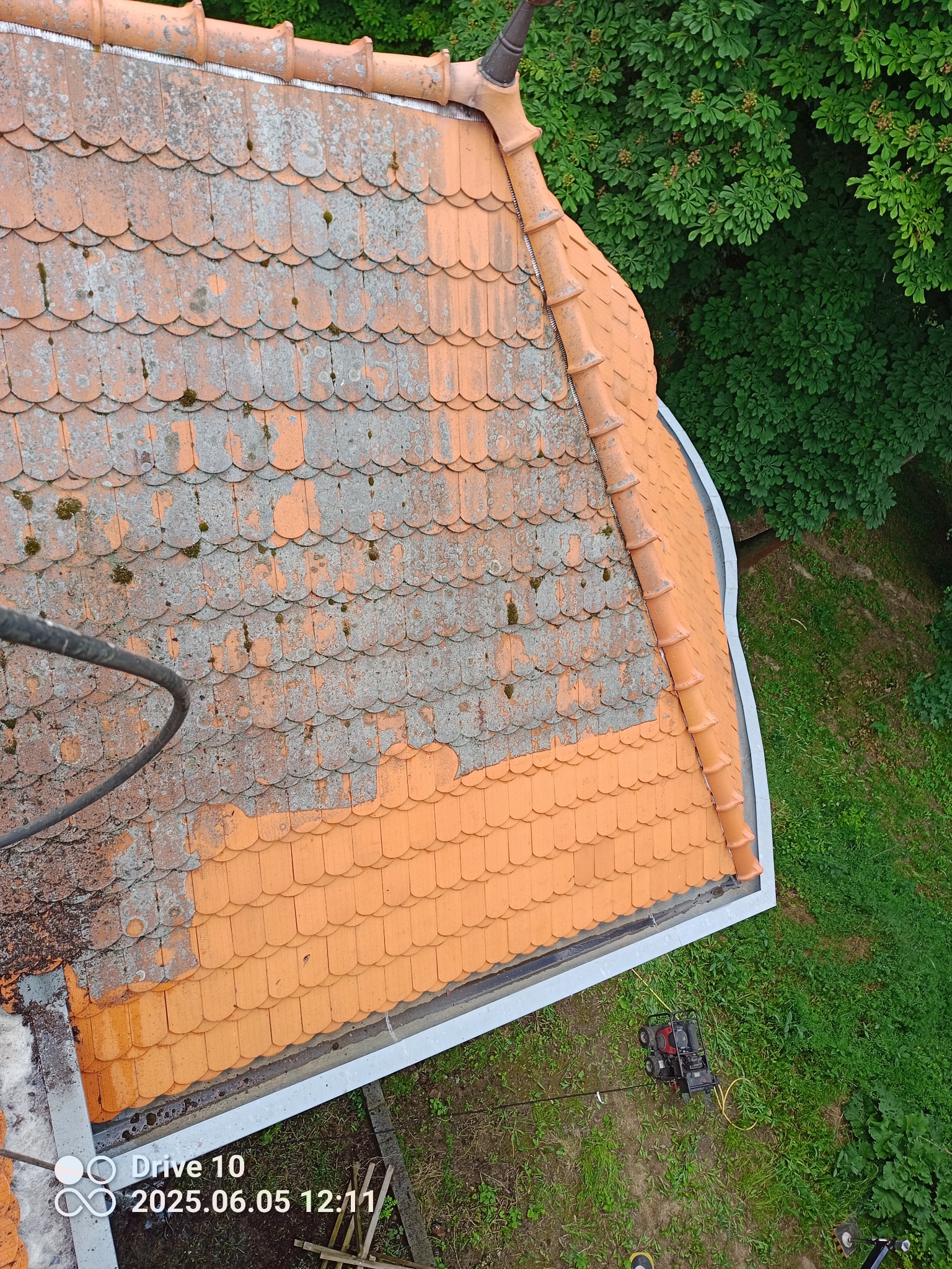 Częściowo wyczyszczony dach z pomarańczowej dachówki karpiówki, widoczne porosty i mech. Na dole sprzęt do czyszczenia pod ciśnieniem i zieleń. Ujęcie z góry.