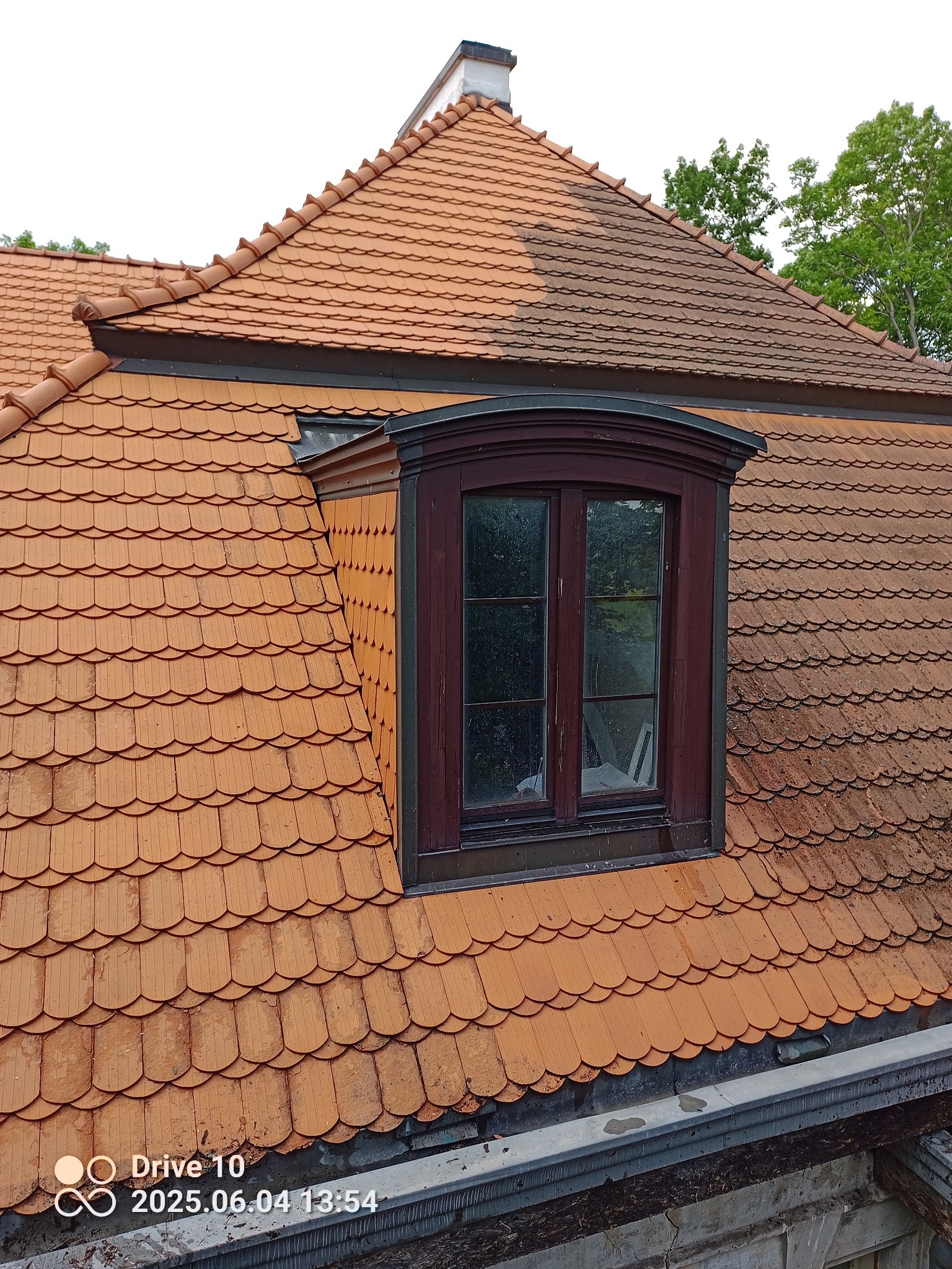 Częściowo wyczyszczony dach z dachówki karpiówki, widoczna lukarna z ciemną ramą okienną. Kontrast czystej i zabrudzonej powierzchni. Ujęcie z dołu, widoczne rynny.