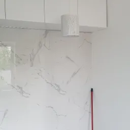 Ściana wyłożona białymi płytkami imitującymi marmur, wisząca biała szafka, lampa z motywem drzewa, mop i śmieciarka wskazują na prace wykończeniowe w łazience.