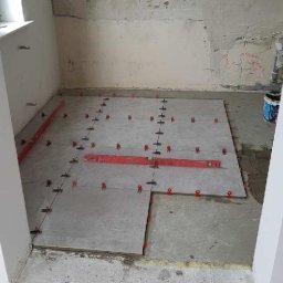 ReM-FACH - Przygotowanie do układania płytek podłogowych w łazience, widoczne poziomice i krzyżyki dystansowe, surowy stan ścian i podłogi.