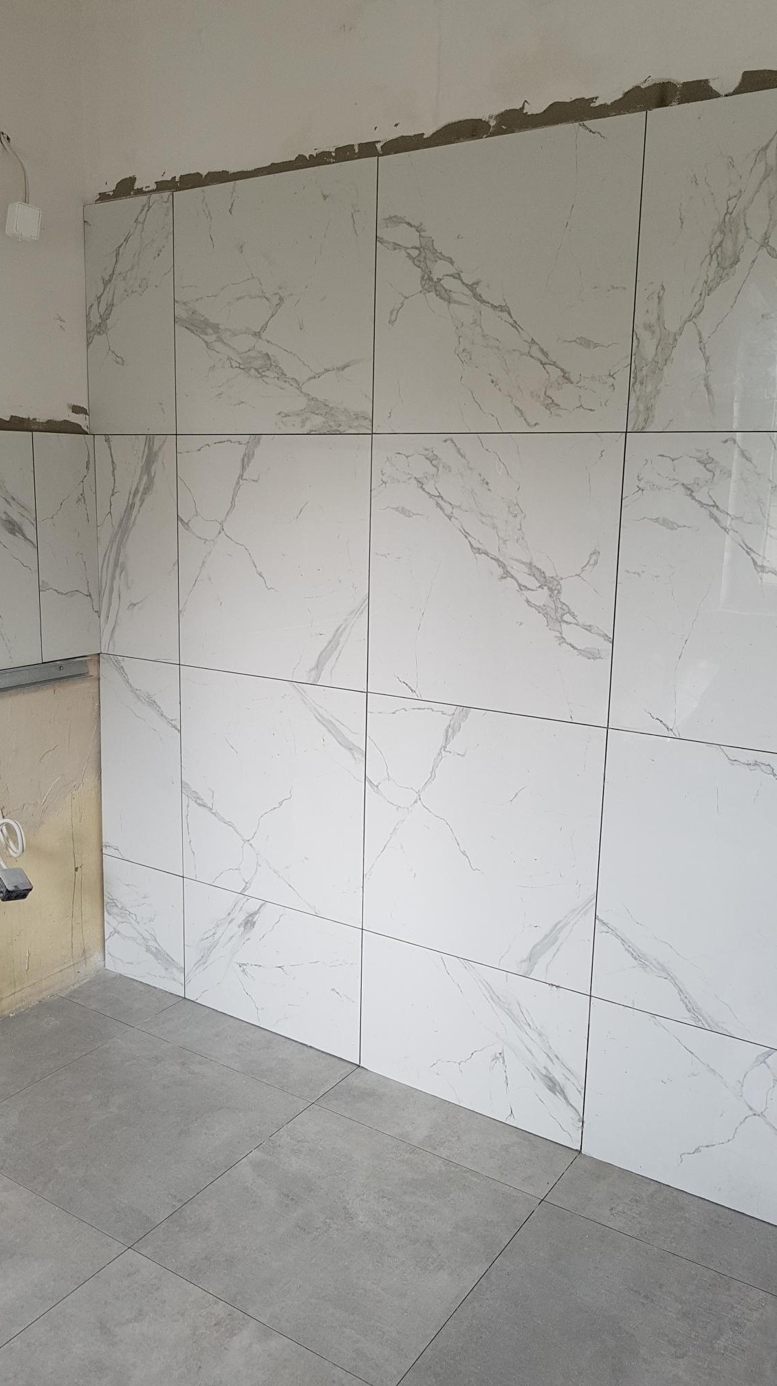 Ściana i podłoga wyłożone płytkami: białe płytki ścienne z szarym marmurkowym wzorem, szare płytki podłogowe, widoczne niedociągnięcia w górnej części ściany.