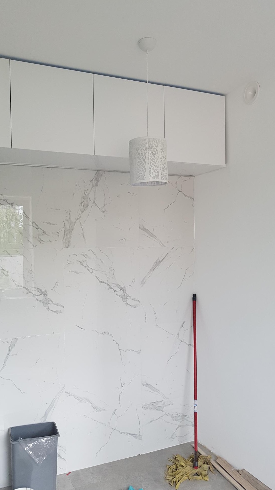 Ściana wyłożona białymi płytkami imitującymi marmur, wisząca biała szafka, lampa z motywem drzewa, mop i śmieciarka wskazują na prace wykończeniowe w łazience.
