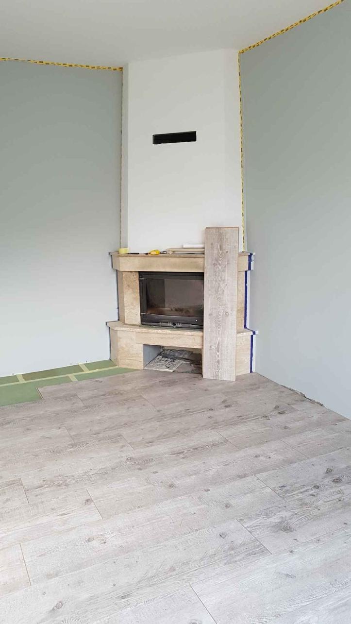 Układanie paneli podłogowych w pokoju z kominkiem, widoczne niedokończone fragmenty podłogi i listwa przypodłogowa zabezpieczona taśmą malarską.