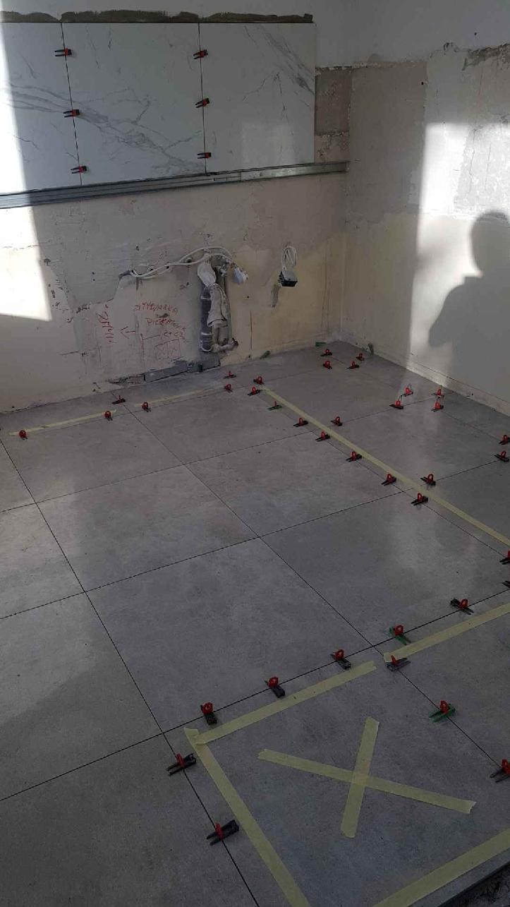 Układanie szarych płytek podłogowych z użyciem klipsów poziomujących i taśmy maskującej w trakcie prac wykończeniowych w Mysłowicach, widoczne także fragmenty płytek ściennych z wzorem marmuru.