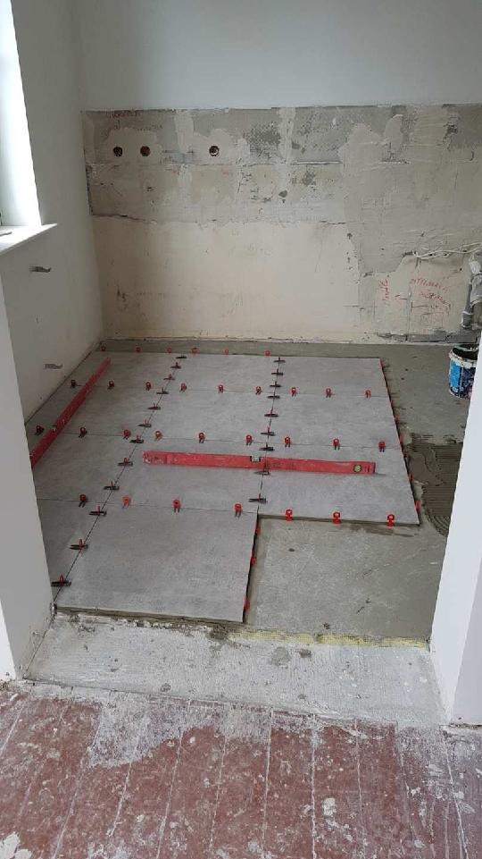 Przygotowanie do układania płytek podłogowych w łazience, widoczne poziomice i krzyżyki dystansowe, surowy stan ścian i podłogi.