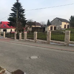 Nowoczesne ogrodzenie z czarnymi, metalowymi przęsłami i słupkami z jasnego kamienia, otaczające posesję z budynkiem o czerwonym dachu, widok z ulicy wyłożonej kostką.