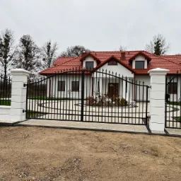 Czarna, kuta brama wjazdowa z białymi, murowanymi słupkami na tle domu z czerwoną dachówką i okrągłym podjazdem z kostki.