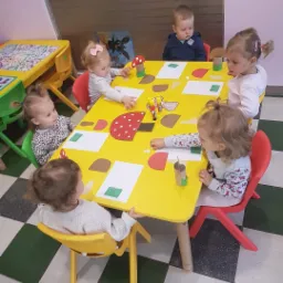 Pięcioro małych dzieci siedzi przy żółtym stoliku, wykonując prace plastyczne z kolorowego papieru i kartonu, tworząc dekoracje w kształcie grzybów i innych elementów.