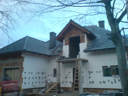 Dom w trakcie budowy z widocznym ociepleniem styropianem, częściowo pokryty ciemną dachówką, z odsłoniętą konstrukcją dachu i kominem. Na pierwszym planie drabina oparta o balkon.